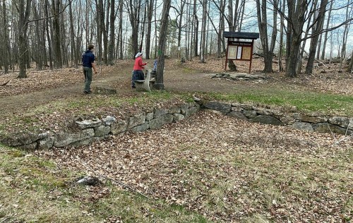 Two people explore a wooded area with a stone wall and a trail sign, surrounded by bare trees and fallen leaves.