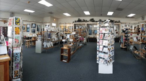 A spacious art gallery filled with framed artworks, greeting cards, and display shelves. Brightly lit and inviting atmosphere.