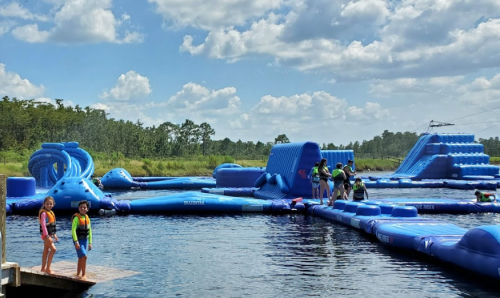 Two children in life jackets stand on a dock, overlooking a blue inflatable water park on a sunny day.