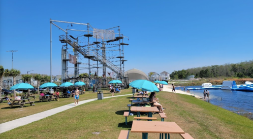 A sunny park with picnic tables and blue umbrellas, featuring a climbing structure and a water area in the background.