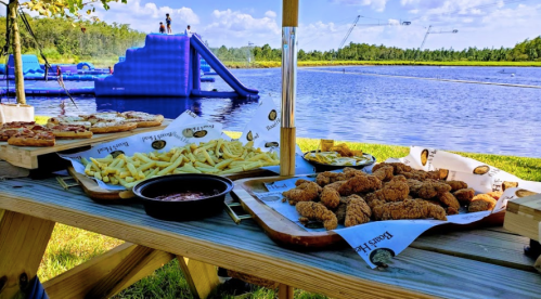 A picnic table with fried chicken, fries, and pizza, overlooking a lake with a water slide and people in the background.