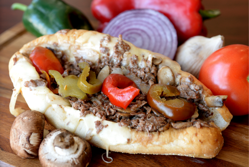 A loaded sandwich with beef, peppers, mushrooms, and onions, surrounded by fresh vegetables on a wooden board.