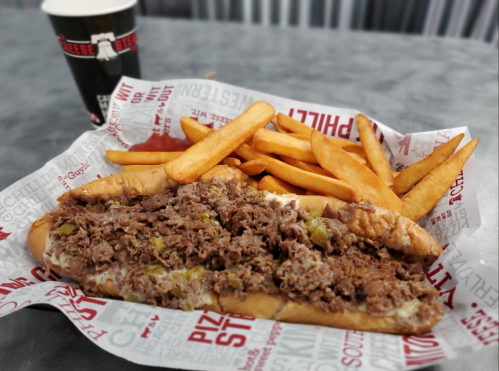 A cheesesteak sandwich with beef and peppers, served with a side of golden fries and a drink in the background.
