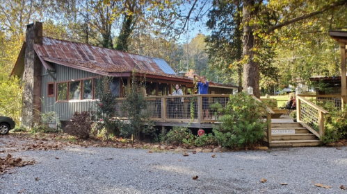 A rustic building with a porch surrounded by trees, featuring two people standing on the deck and a gravel parking area.