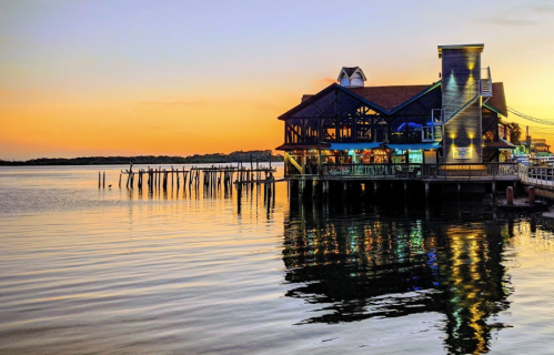 A waterfront restaurant at sunset, reflecting colorful lights on calm water with a pier in the background.