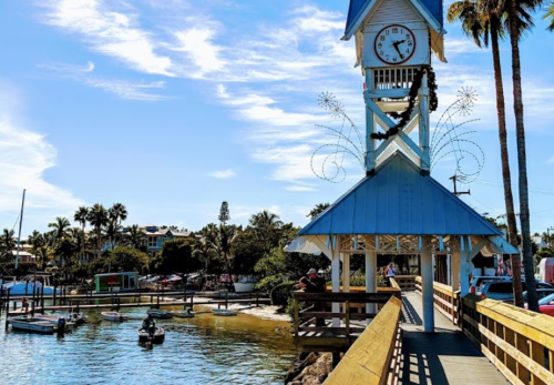 A clock tower with blue accents overlooks a calm waterway, surrounded by palm trees and boats on a sunny day.