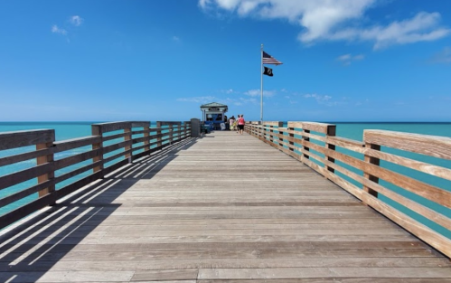 A wooden pier extends over turquoise waters, with people walking and an American flag waving in the background.