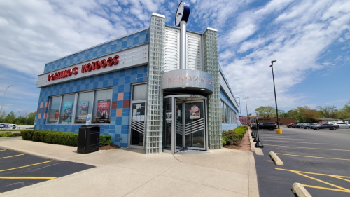 Exterior of a colorful hot dog restaurant with a glass entrance and a clear blue sky in the background.
