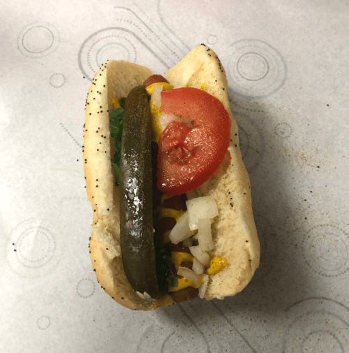 A hot dog in a bun topped with mustard, diced onions, pickles, tomato, and green relish on a patterned paper background.