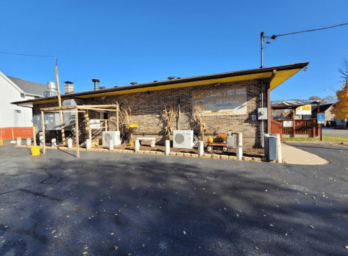 A brick building with a sign for hot dogs, featuring outdoor seating and seasonal decorations. Clear blue sky above.