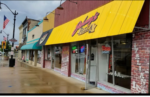 A pizza shop with a yellow awning and neon signs, located on a rainy street with colorful storefronts nearby.