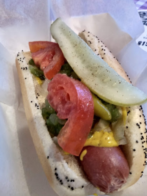 A close-up of a hot dog in a poppy seed bun, topped with mustard, tomatoes, pickles, and green relish.