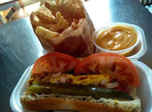 A hot dog topped with tomatoes, onions, and pickles, served with crinkle-cut fries and a cup of cheese sauce.