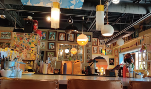 A cozy café interior with colorful decor, framed art on the walls, and warm lighting hanging from the ceiling.