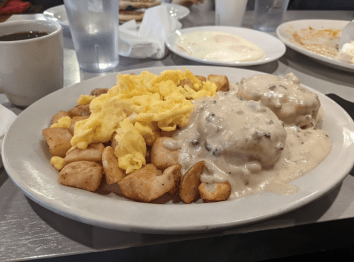 A plate of scrambled eggs, biscuits with gravy, and seasoned potatoes, accompanied by a cup of coffee.