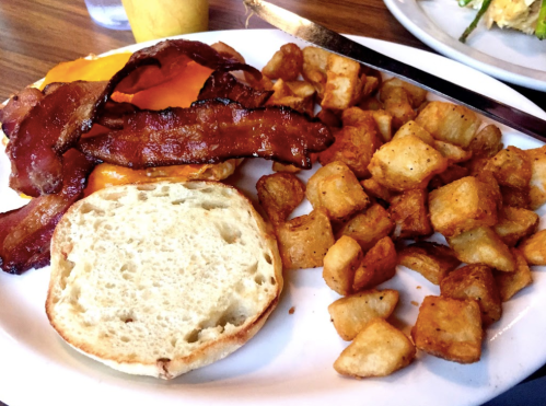 A plate of breakfast featuring crispy bacon, a cheese-topped egg, golden potatoes, and a toasted English muffin.