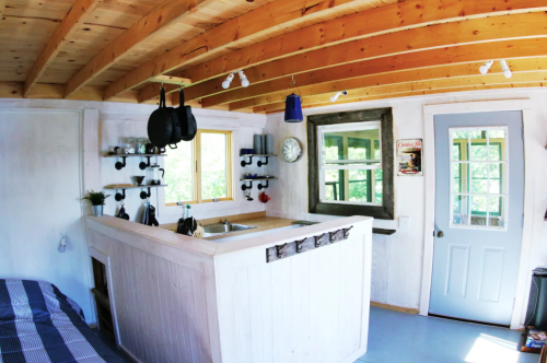 Cozy interior of a small cabin with wooden beams, a kitchenette, and large windows letting in natural light.