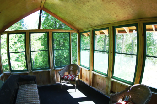 A bright, airy sunroom with large windows, wooden walls, and cozy seating, surrounded by lush greenery outside.