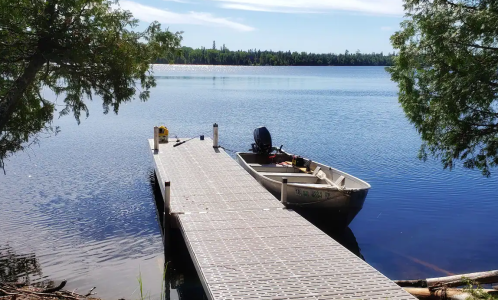 A calm lake scene featuring a wooden dock and a small boat moored beside it, surrounded by trees and clear blue skies.