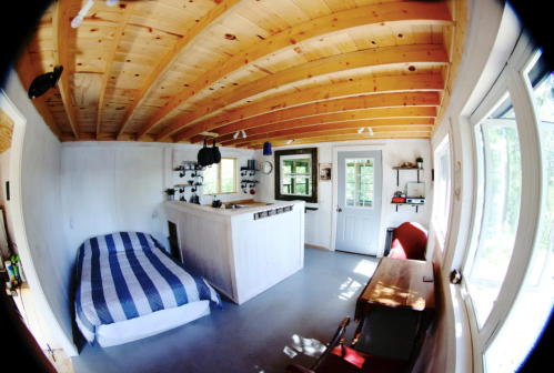 Cozy interior of a cabin featuring a bed, wooden ceiling, small kitchen area, and a table with chairs.