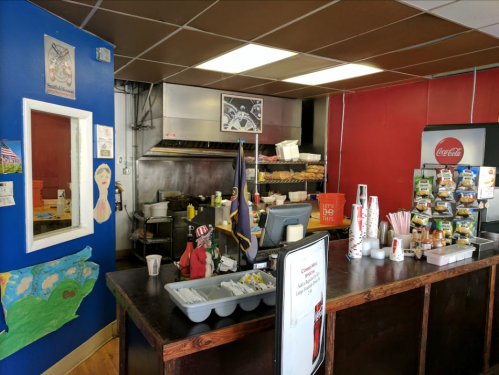 A small diner interior with a counter, kitchen area, and colorful wall decorations, including a mirror and Coca-Cola products.