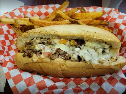 A cheesesteak sandwich with melted cheese and toppings, served with golden-brown fries in a red-checkered basket.