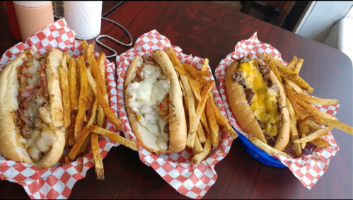 Three loaded hot dogs with toppings, served with crispy fries, on a checkered tablecloth.