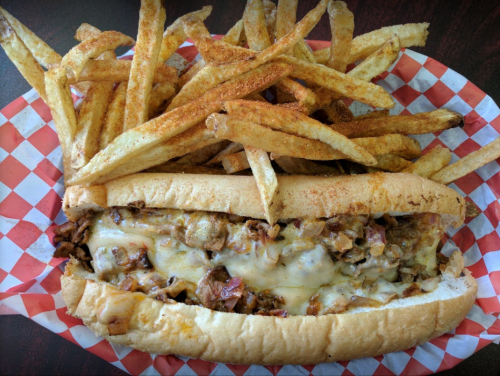 A loaded sandwich with melted cheese and meat, served with a side of crispy, seasoned French fries in a red-checkered basket.