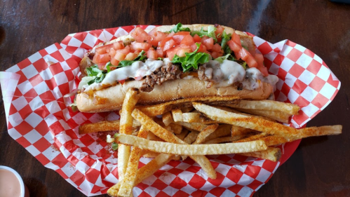 A loaded sandwich with meat, lettuce, and tomatoes, served with a side of crispy fries in a red-checkered basket.