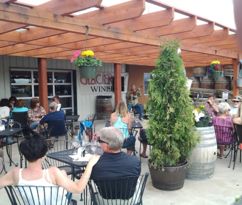 A lively outdoor winery scene with people seated at tables, enjoying music and drinks under a wooden pergola.