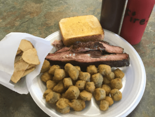 A plate with sliced brisket, fried okra, potato chips, and a slice of toast, accompanied by two bottles of sauce.