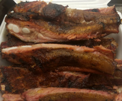A close-up of several pieces of grilled, smoky ribs stacked in a white takeout container.