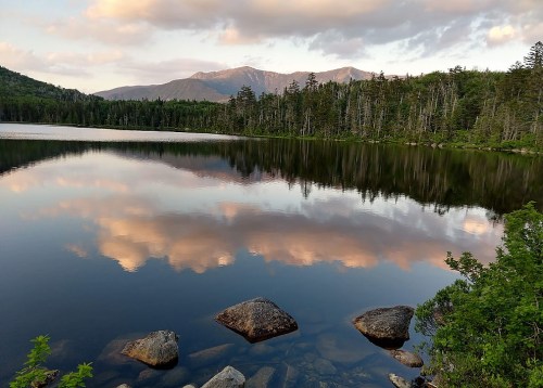 A serene lake reflects mountains and clouds, surrounded by lush trees and rocky shores under a soft sky.