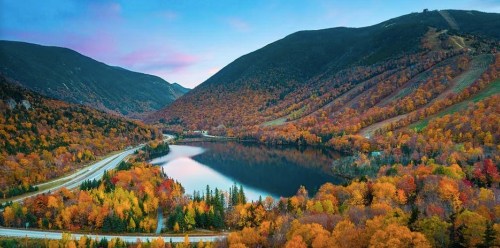 A scenic view of a lake surrounded by vibrant autumn foliage and rolling hills under a colorful sky.