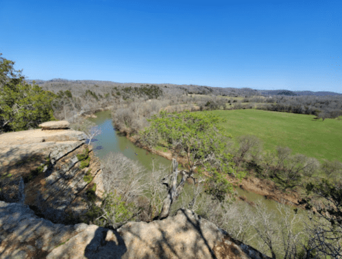 A scenic view from a rocky cliff overlooking a winding river and lush green fields under a clear blue sky.