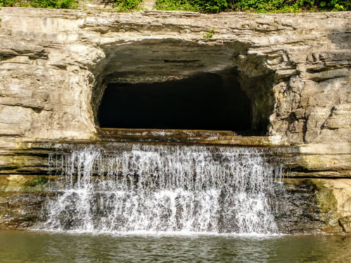 A small waterfall cascades over rocky terrain into a dark cave entrance surrounded by greenery.
