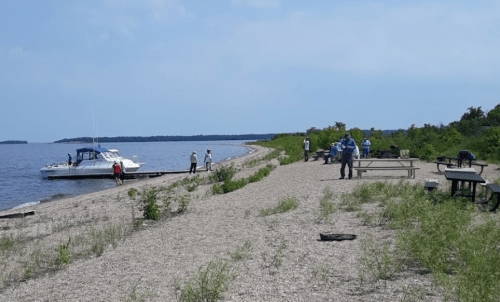 A sandy shoreline with a boat docked nearby, people walking, and picnic tables under a clear blue sky.