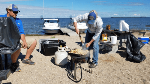 A person cooks on a portable stove by the water while another sits nearby, with boats in the background.