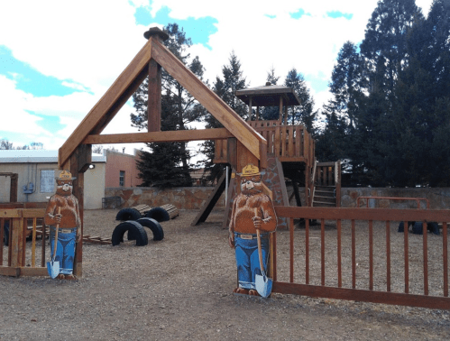 Wooden playground with a climbing structure, surrounded by trees, featuring cartoon bear cutouts at the entrance.