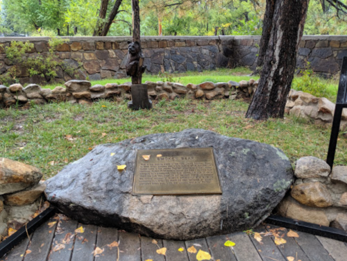A large rock with a plaque sits in a grassy area surrounded by trees and a stone wall, with a wooden sculpture nearby.
