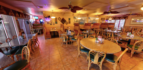A spacious restaurant interior with round tables, rattan chairs, and tropical decor, featuring warm lighting and booths.
