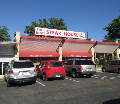 Exterior of "The Best Steak House" with red awnings and parked cars in front under a clear blue sky.