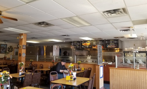 A cozy diner interior with tables, a menu board, and a lone customer reading at a table. Flowers adorn the tables.