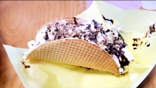 A waffle taco filled with creamy dessert, topped with crushed nuts and chocolate drizzle, resting on yellow paper.