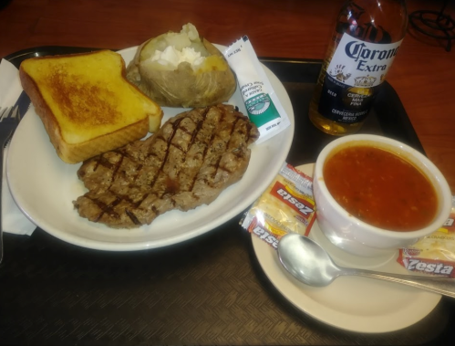 A plate with grilled steak, baked potato, toast, a bowl of soup, and a bottle of Corona beer on a tray.