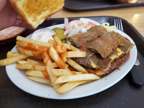 A plate with a burger topped with sliced meat, fries, pickles, tomatoes, and a hand holding a piece of toast.