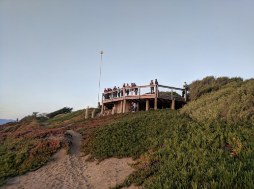A wooden observation deck on a hillside with people enjoying the view, surrounded by greenery and a clear sky.