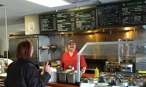 A smiling employee in a red shirt stands behind a counter, with a menu board and food items visible. A customer gestures.