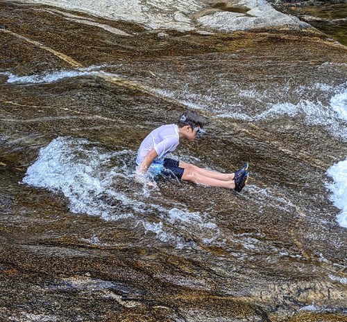 A child sits on a rocky surface in a stream, splashing water around while enjoying a sunny day outdoors.