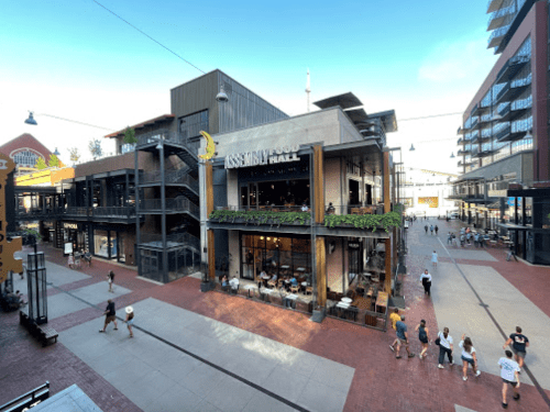 A bustling outdoor shopping area featuring Assembly Food Hall, with people walking and modern buildings in the background.
